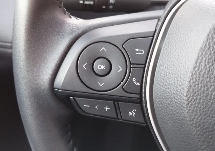 Gros plan sur les commandes gauche du volant gris du Toyota Corolla Cross 1.8 140ch Dynamic 2025, boutons multifonctions visibles.