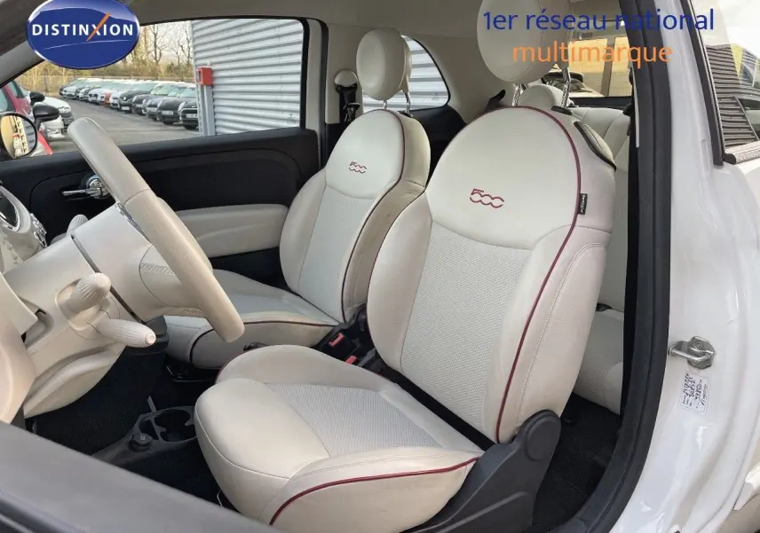 Vue intérieure côté conducteur de la Fiat 500 blanche 1.0 Hybrid 70ch DolceVita avec sièges cuir blanc et surpiqûres rouges.