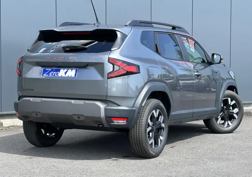 Vue 3/4 arrière droite du Dacia Duster 2025 gris schiste avec jantes alliage et barres de toit noires.