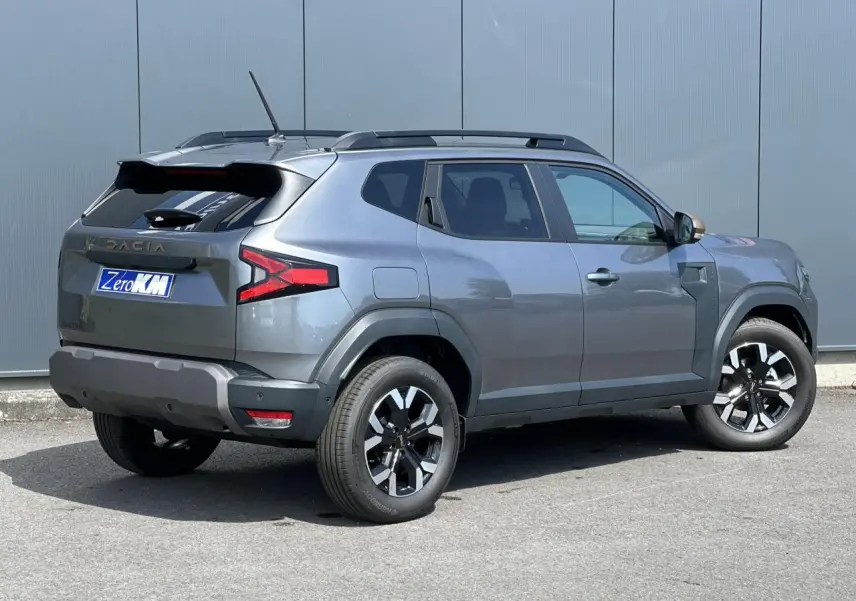 Dacia Duster Gris Schiste en 3/4 arrière droit, avec barres de toit noires et jantes alliage 17 pouces.