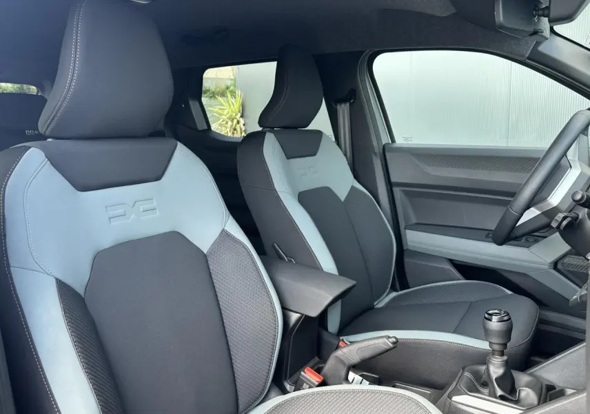 Vue intérieure côté conducteur du Dacia Duster 2025, sièges bi-ton gris clair et foncé avec logo embossé sur appuie-tête.