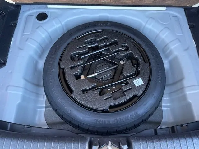 Vue en plongée sur la roue de secours noire et ses outils rangés dans le coffre argent lunaire d'une Kia CEE'D 2024.