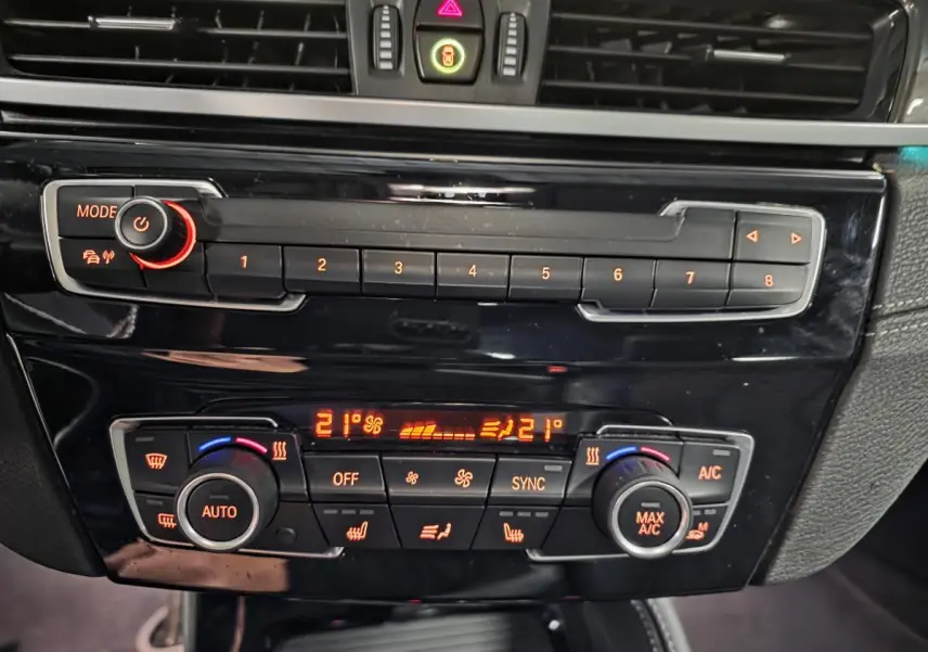 Gros plan sur la console centrale noire brillante du BMW X1 blanc, avec commandes climatisation bi-zone et boutons rétroéclairés orange.