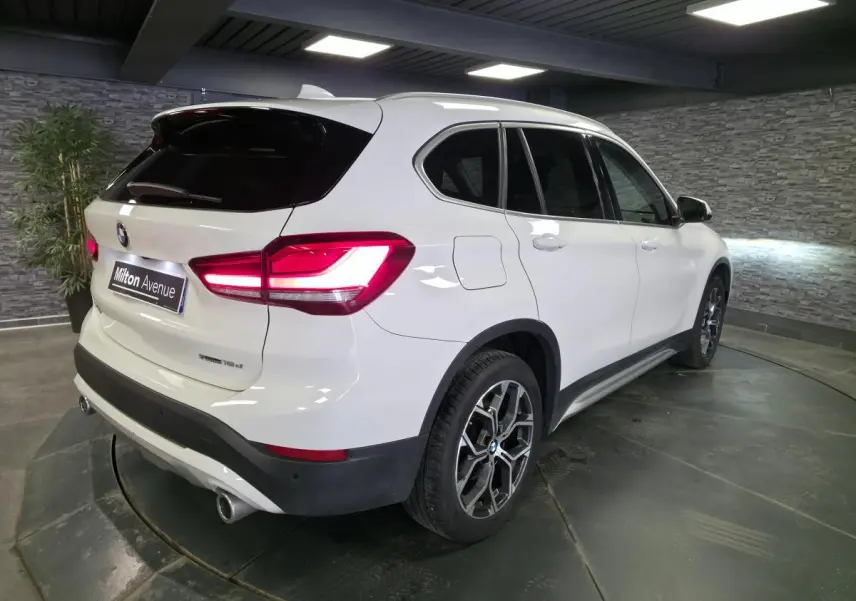 BMW X1 sDrive 18d blanc vue 3/4 arrière droit, avec jantes alliage 18 pouces et sorties d’échappement chromées.