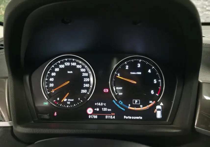 Gros plan sur le tableau de bord numérique du BMW X1 blanc, affichant la vitesse, le régime moteur et les alertes.