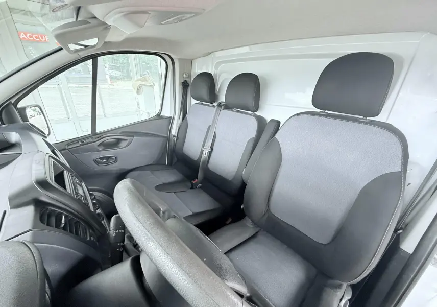 Vue intérieure côté conducteur de l'Opel Vivaro blanc, montrant les sièges tissu gris et noir et le tableau de bord.