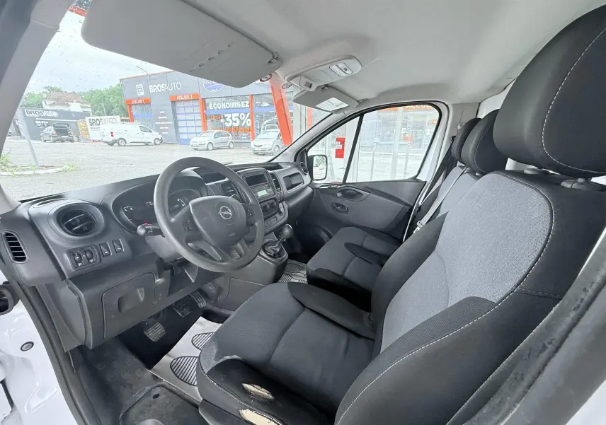 Vue intérieure côté conducteur de l'Opel Vivaro blanc 2018, sièges tissu gris noir et tableau de bord noir.