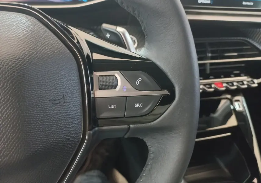 Gros plan sur le volant cuir noir de la Peugeot 208 2022 avec commandes multimédia et tableau de bord tactile en arrière-plan.
