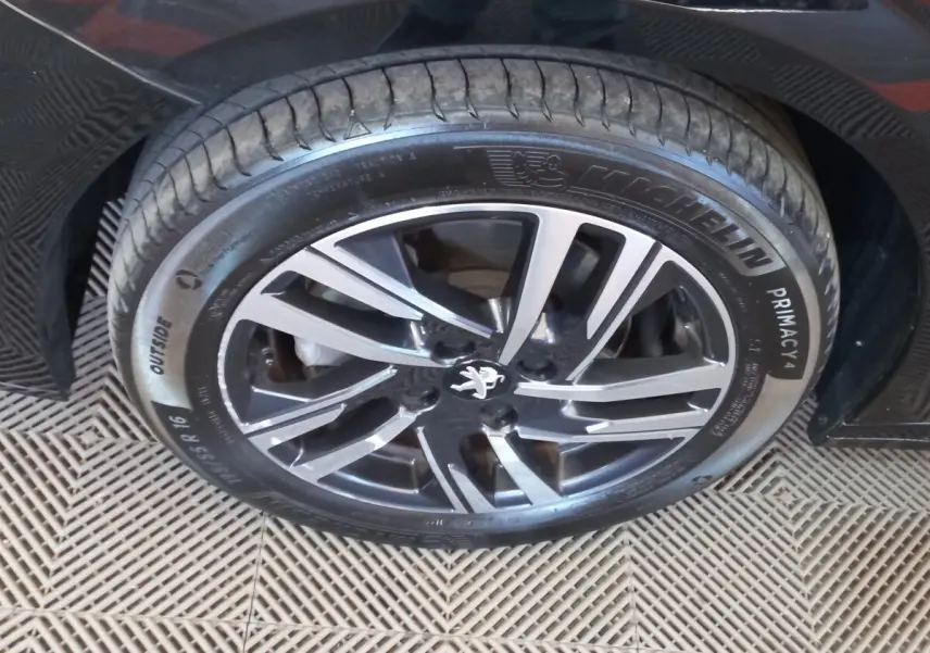 Gros plan sur la jante alliage noire et argentée avec logo Peugeot, montée sur un pneu Michelin Primacy 4, voiture noire visible en arrière-plan.