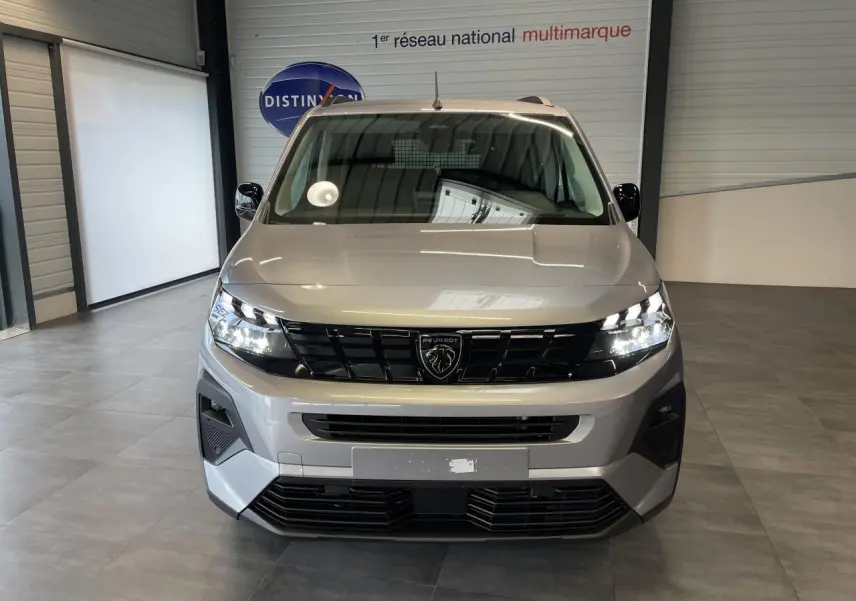 Vue de face du Peugeot Rifter gris Artense avec calandre noire et signature lumineuse LED allumée en intérieur showroom.