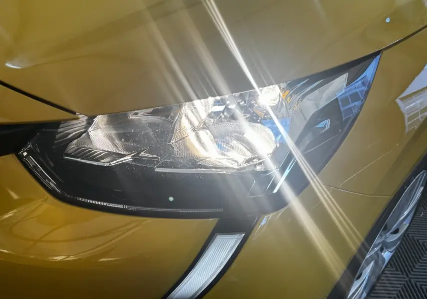 Gros plan sur le phare avant droit d’une Peugeot 208 jaune 2023 avec reflets lumineux sur la carrosserie.