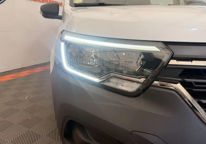 Gros plan sur le phare avant gauche allumé d’un Renault Kangoo blanc, soulignant la calandre noire et chromée.