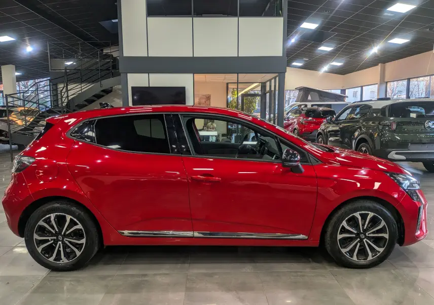 Profil latéral droit d'une Renault Clio V rouge flamme en showroom, avec jantes alliage et détails chromés sur les bas de porte.