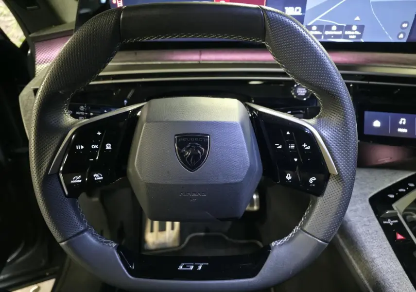 Gros plan sur le volant cuir perforé noir avec badge GT du Peugeot 3008 1.2i Hybrid 2025, tableau de bord numérique en arrière-plan.