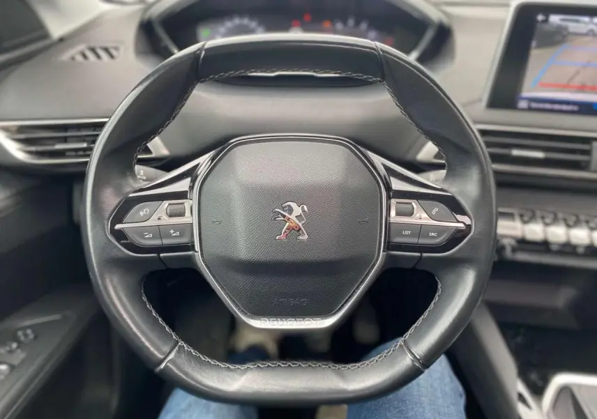 Vue rapprochée du volant compact noir du Peugeot 3008 2019 avec commandes intégrées et tableau de bord flou en arrière-plan.