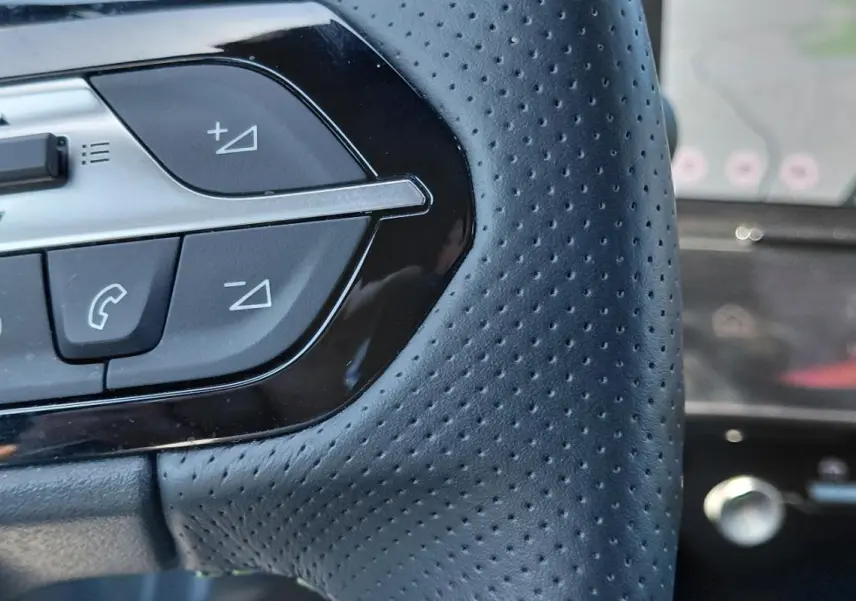 Gros plan sur les commandes gauche du volant cuir perforé d'une Peugeot 308 Hybrid 145 e-DCS6 GT gris, avec cockpit numérique en arrière-plan.