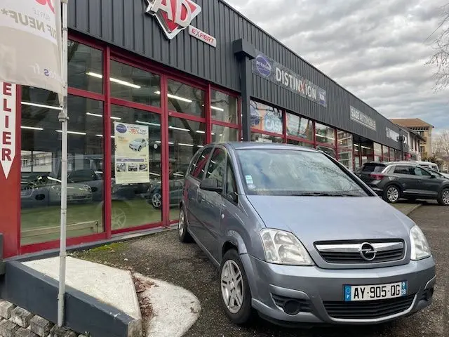 Opel Meriva grise vue 3/4 avant droit stationnée devant un garage avec façade vitrée et enseigne AD Expert.