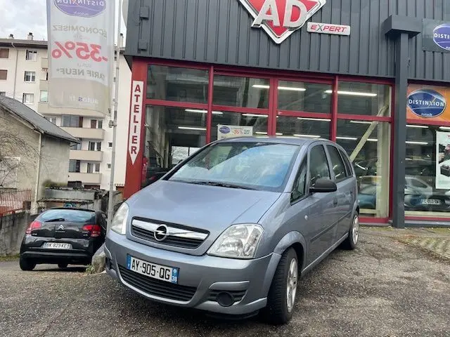 Opel Meriva grise vue 3/4 avant droit devant un garage AD Expert avec ses phares et calandre visibles.