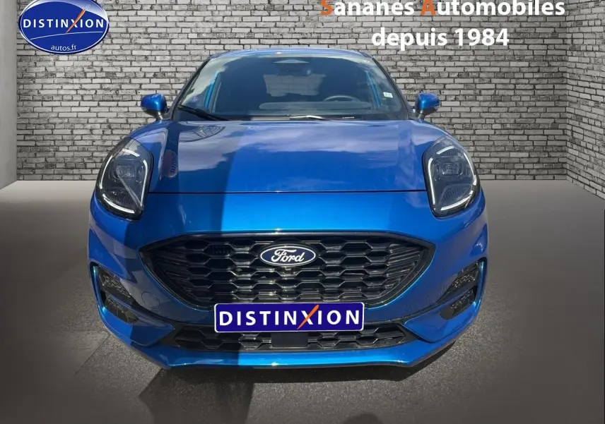 Vue frontale d'une Ford Puma bleu métallisé avec calandre noire et phares LED, sur fond de mur en briques grises.