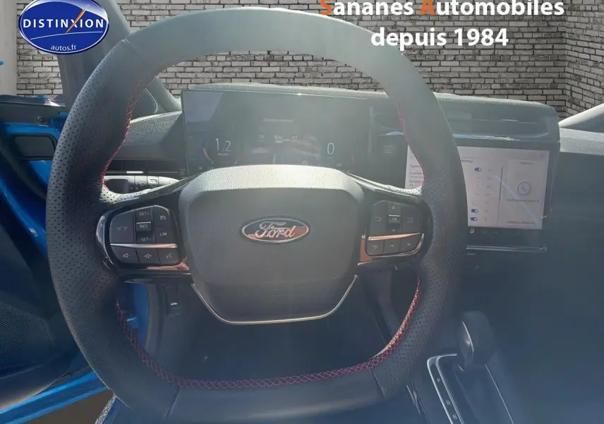 Vue intérieure centrée sur le volant perforé avec surpiqûres rouges du Ford Puma bleu 2025, tableau de bord numérique et écran tactile visibles.