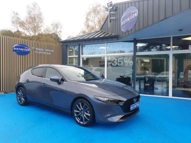 Vue 3/4 avant gauche de la Mazda3 2024 Platinum Quartz Métallisé garée devant un showroom Distinxion.