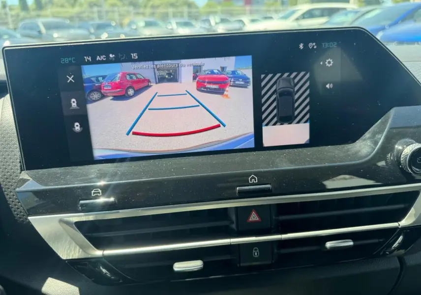 Écran intérieur montrant la caméra de recul avec vue arrière sur un parking ensoleillé.