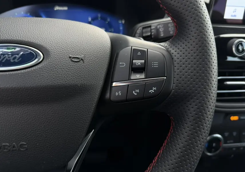 Gros plan sur le volant perforé noir avec surpiqûres rouges du Ford Kuga 2023, boutons de commande visibles à droite.