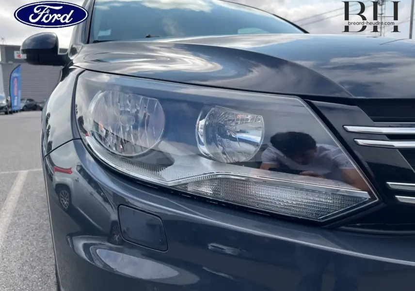 Gros plan sur le phare avant gauche d’un Volkswagen Tiguan gris poivre avec calandre chromée visible.