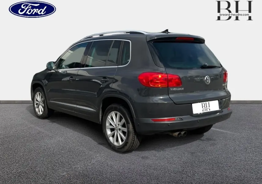 Vue 3/4 arrière droite d'un Volkswagen Tiguan gris poivre avec jantes alu et barres de toit visibles.
