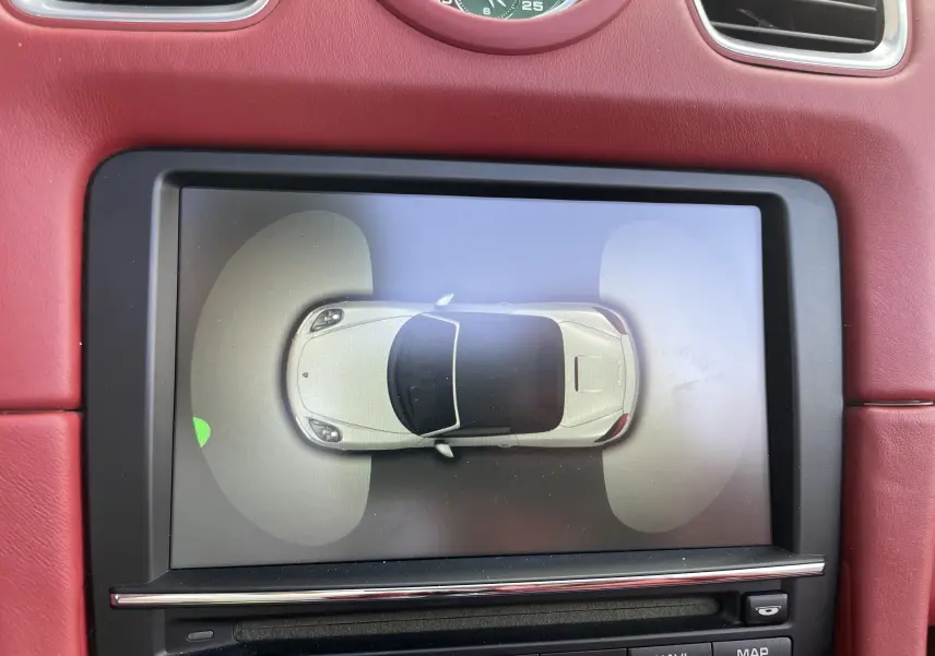 Affichage intérieur du système d'assistance parking montrant une Porsche Boxster S 981 vue de dessus en blanc sur écran intégré au tableau de bord rouge grenat.
