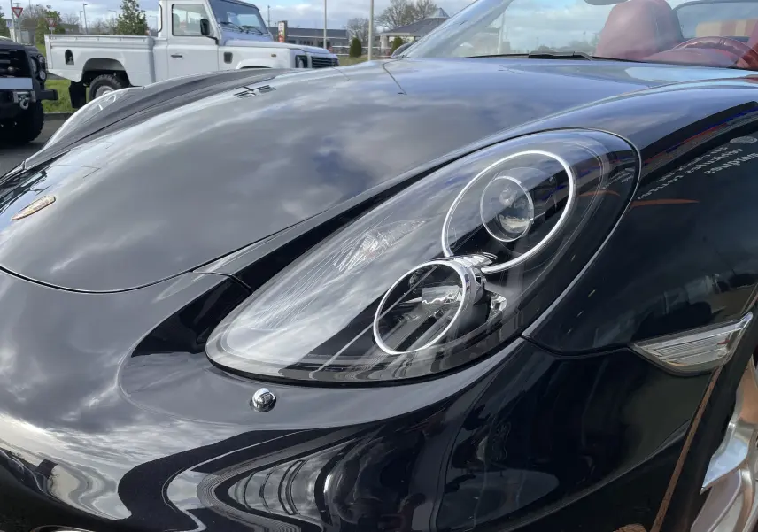 Gros plan sur le phare avant droit d'une Porsche Boxster S 981 noire intense avec intérieur cuir rouge grenat visible.