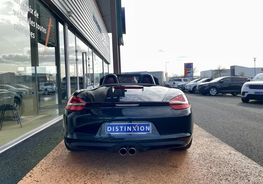Vue arrière d'une Porsche Boxster S noire intense cabriolet, garée devant un showroom avec intérieur cuir rouge visible.