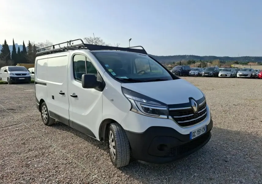 Fourgon Renault Trafic blanc vu en 3/4 avant droit avec galerie de toit, stationné sur un sol gravillonné.