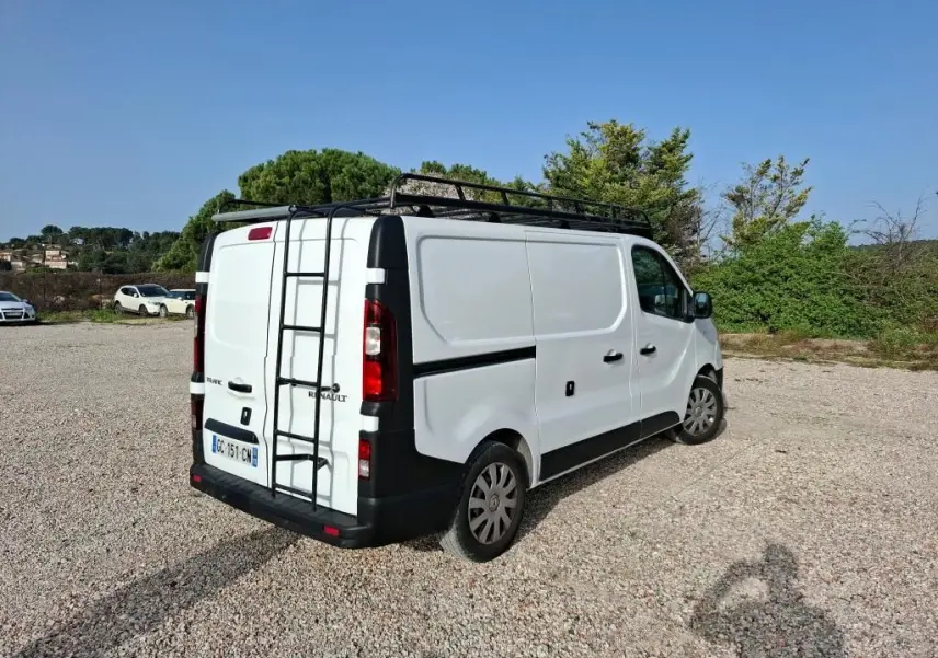 Renault Trafic Fourgon blanc vu en 3/4 arrière droit avec galerie et échelle sur le toit, stationné sur un sol gravillonné.