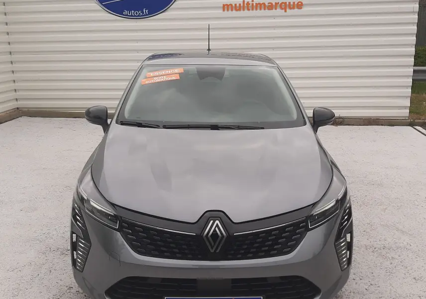 Vue avant d'une Renault Clio V gris schiste avec calandre noire et phares LED, stationnée devant un mur publicitaire.