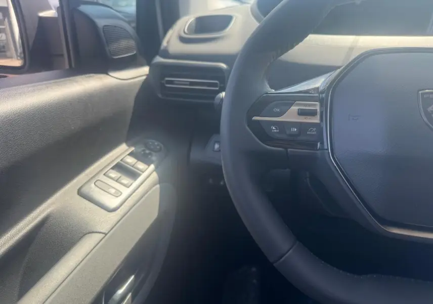 Vue intérieure côté conducteur du Peugeot Rifter gris 2026, volant multifonctions et commandes des vitres visibles.