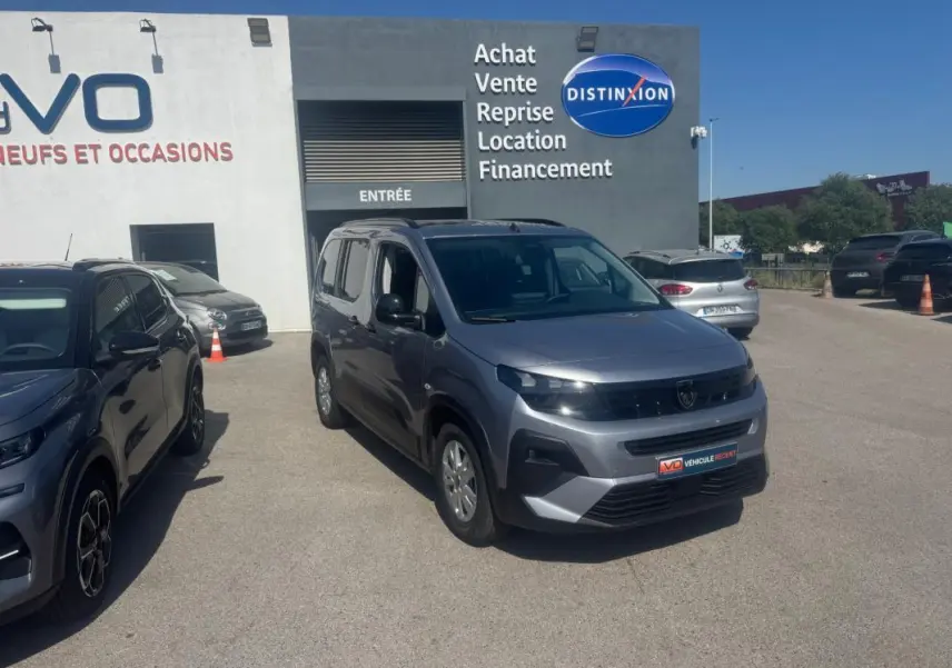Peugeot Rifter gris vue 3/4 avant droit, avec barres de toit et jantes alliage, stationné devant un bâtiment commercial.