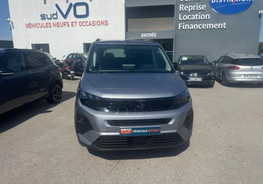 Vue avant d’un Peugeot Rifter gris 2026 avec calandre noire et logo Peugeot visible, stationné devant un concessionnaire.
