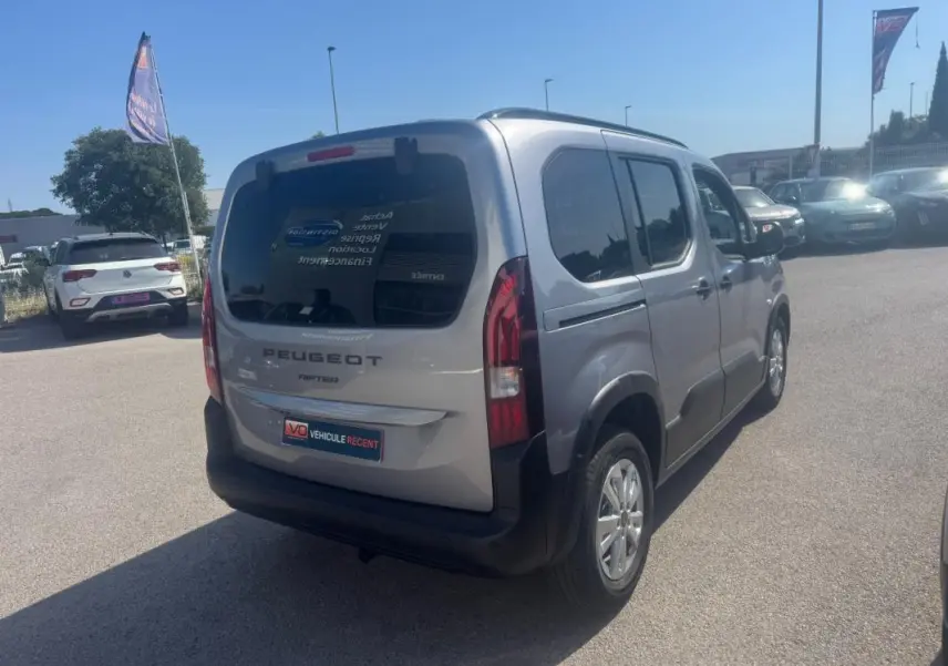 Peugeot Rifter gris vue 3/4 arrière droit, avec barres de toit et vitres teintées, stationné en extérieur.