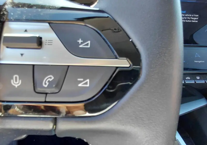 Gros plan sur les commandes au volant noir du Peugeot 408 Hybrid 145 e-DCS6 Allure 2025, avec écran tactile en arrière-plan.