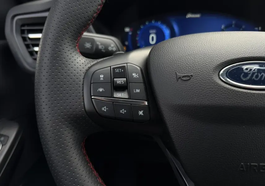 Gros plan sur le volant perforé noir avec surpiqûres rouges du Ford Kuga 2023, commandes intégrées et tableau de bord numérique bleu en arrière-plan.