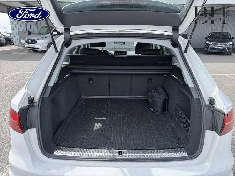 Coffre ouvert vu de l'arrière d'une Audi A4 Avant blanc glacier métallisé, avec tapis de coffre noir et filet latéral.