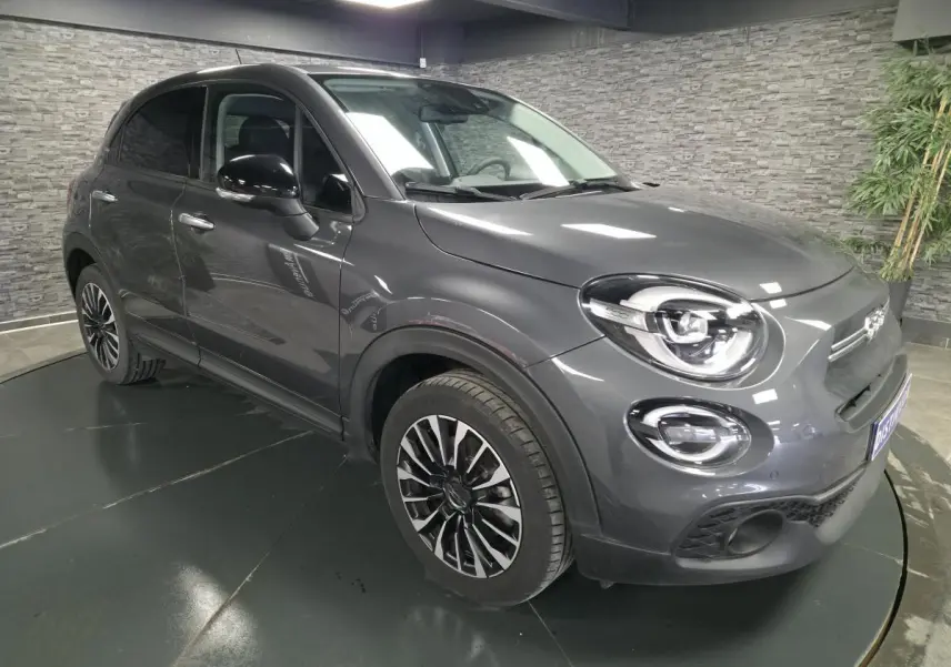 Fiat 500X gris vue 3/4 avant droit, jantes alliage 17'', phares LED et rétroviseurs noirs dans un intérieur moderne.
