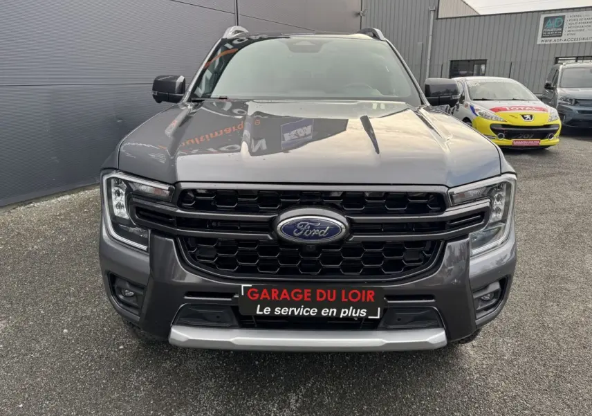 Vue avant d'un Ford Ranger gris métallisé avec calandre noire et logo Ford visible, stationné devant un mur gris.