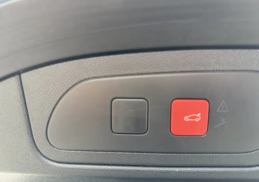 Boutons de commande du coffre, dont un rouge avec pictogramme de voiture, sur panneau intérieur noir texturé.