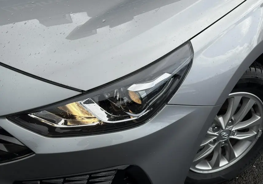 Gros plan sur le phare avant droit et la jante alliage d'une Hyundai i30 SW T-GDI 120 Modern gris argent sous la pluie.