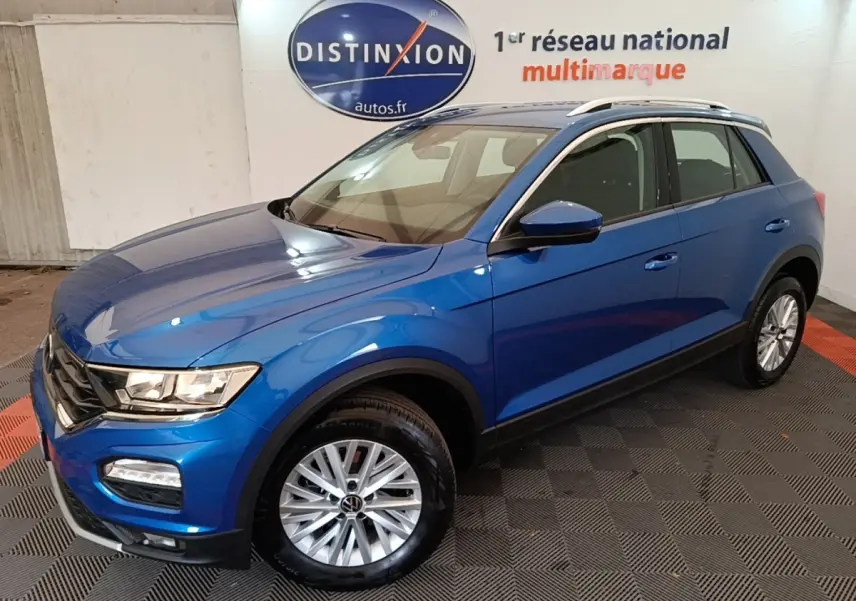 Volkswagen T-Roc bleu en 3/4 avant droit, avec jantes argentées et barres de toit blanches visibles en intérieur showroom.
