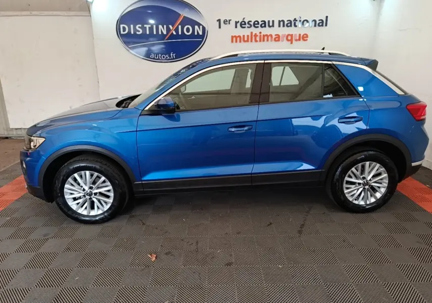 Vue latérale droite du Volkswagen T-Roc bleu 2020, avec jantes alliage et barres de toit noires visibles en intérieur showroom.