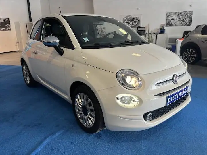 Vue 3/4 avant d'une FIAT 500 blanche avec phares allumés, sur tapis bleu en showroom.