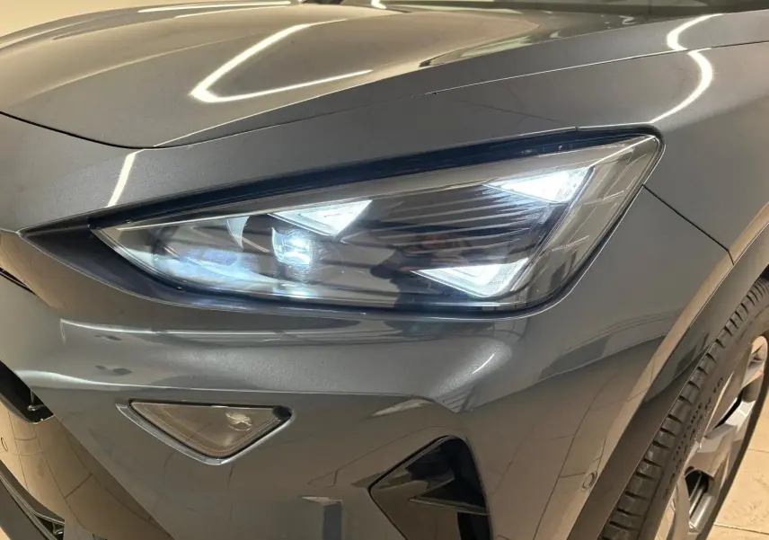 Gros plan sur le phare avant droit allumé d'un CUPRA Formentor gris magnétique avec détails du pare-chocs et roue.